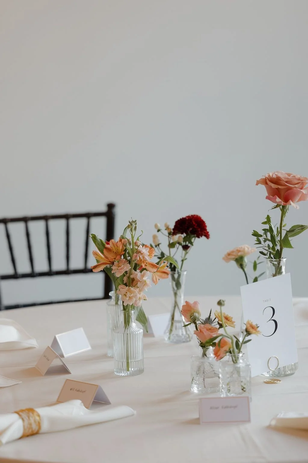 Cluster of bud vases with coral and terracotta blooms beside a table number on a reception table