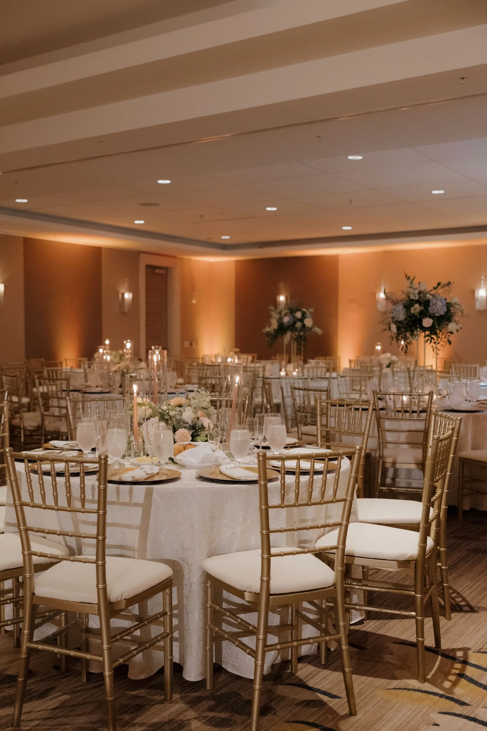 Reception ballroom with round tables, gold chiavari chairs, tall floral centerpieces, and warm uplighting