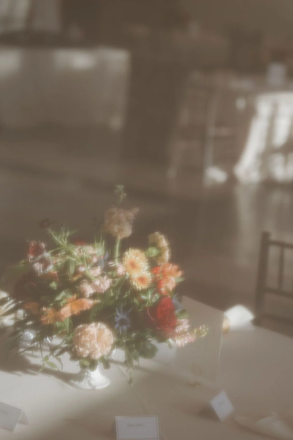 Low centerpiece of peach, terracotta, and blue blooms backlit on a reception table
