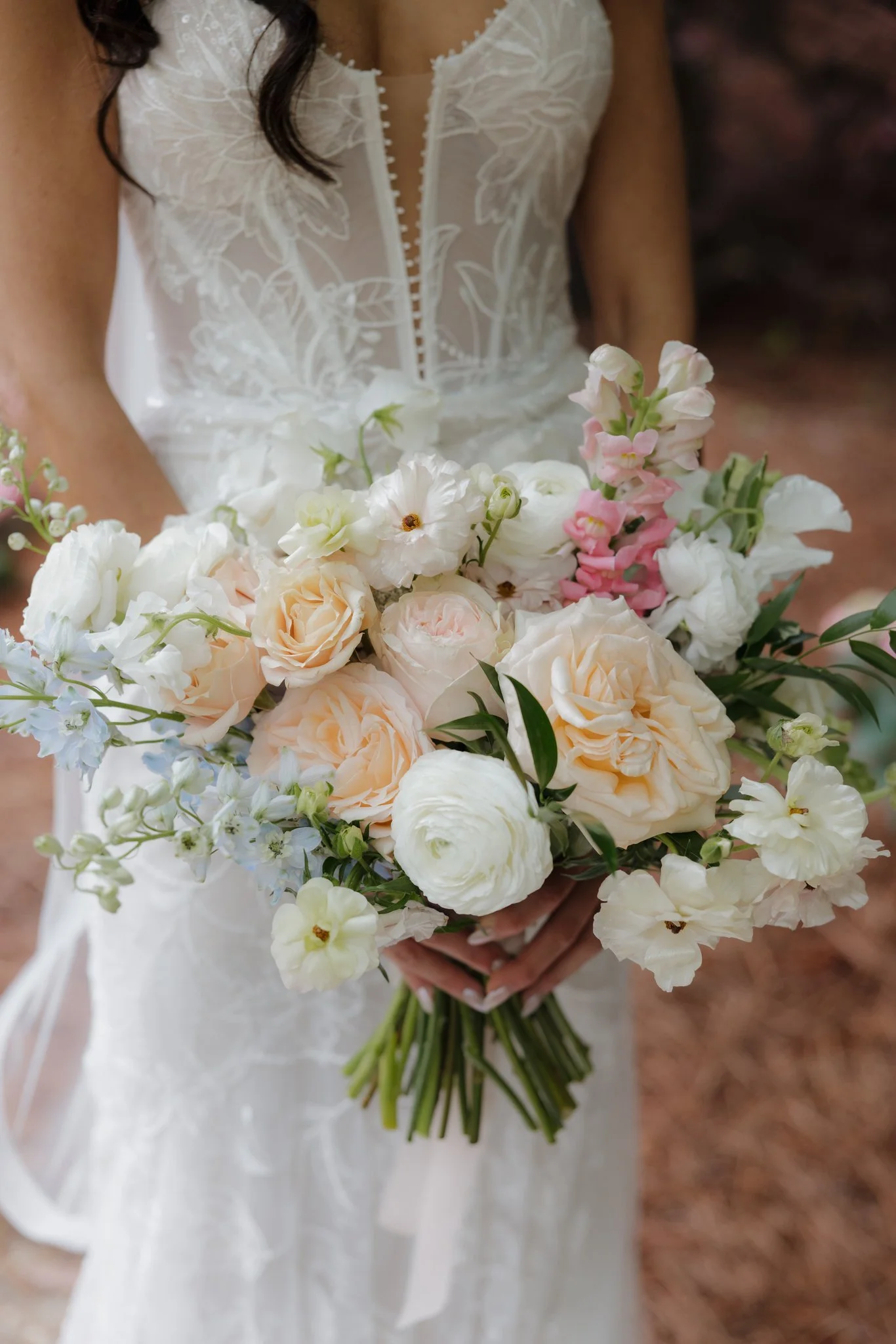 Bride holding a romantic pastel wedding bouquet with blush garden roses, white ranunculus, delphinium, and soft pink blooms, paired with a lace wedding dress featuring a sheer bodice and floral detailing.