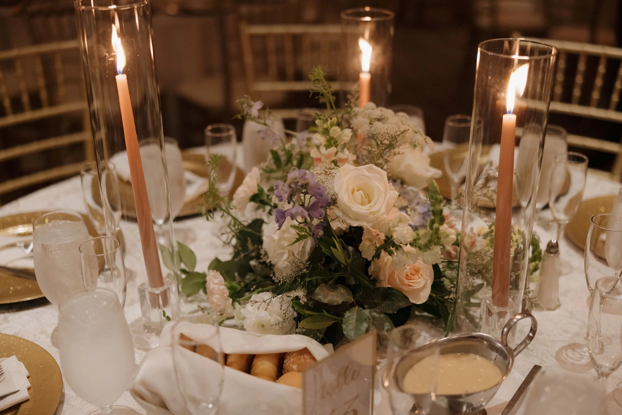 Round reception table with a low centerpiece of white roses and lavender blooms, tall taper candles, and gold chargers