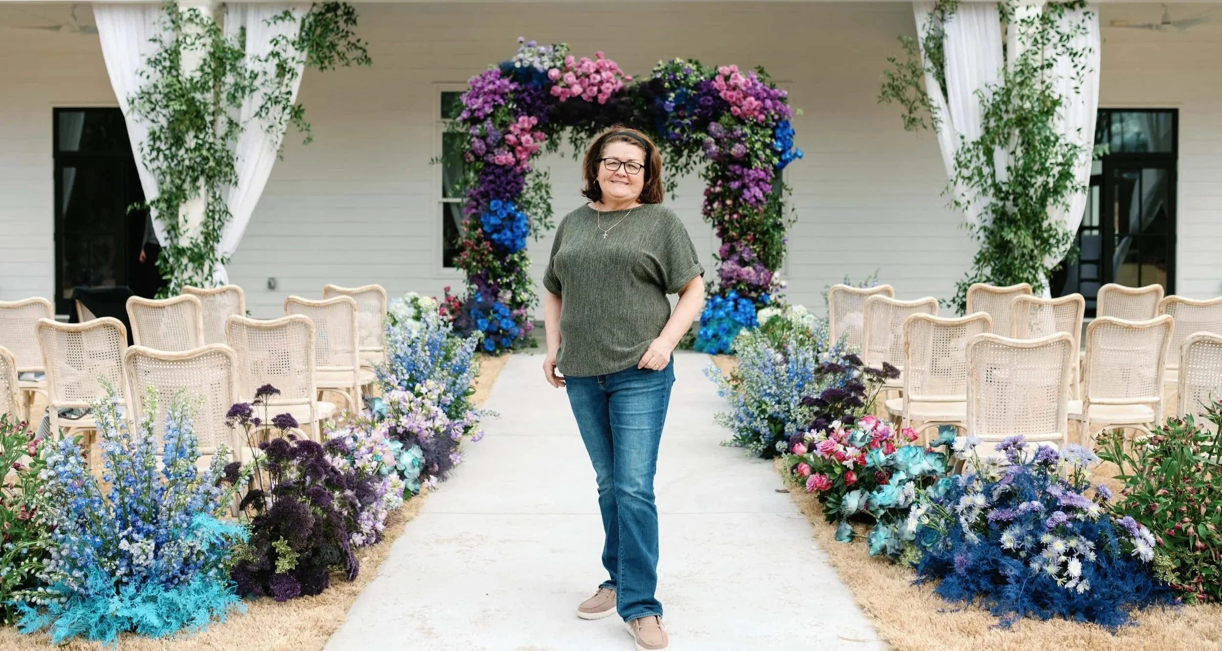 Lead floral designer standing confidently in front of a colorful ceremony floral arch of purple, pink, and blue flowers at an outdoor wedding venue.
