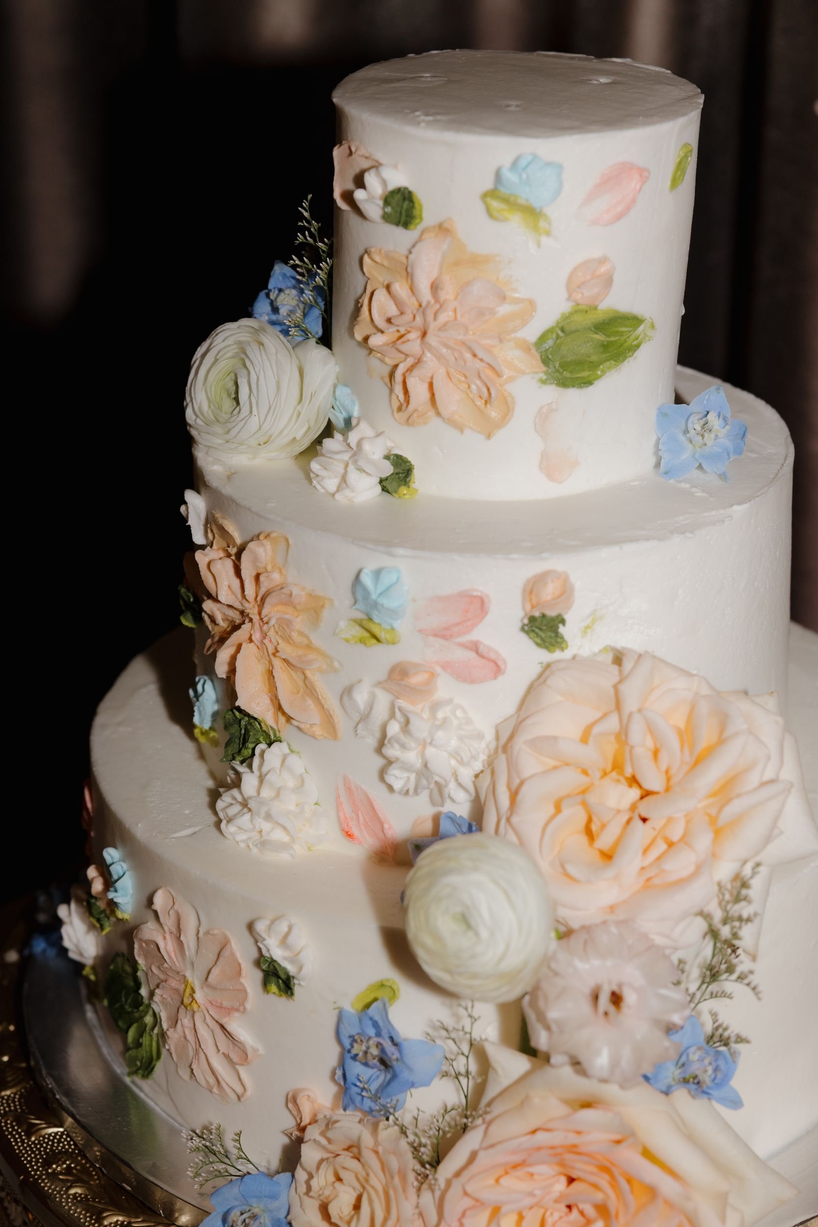 Three-tier white wedding cake with hand-painted florals and fresh peach roses, white ranunculus, and blue blooms