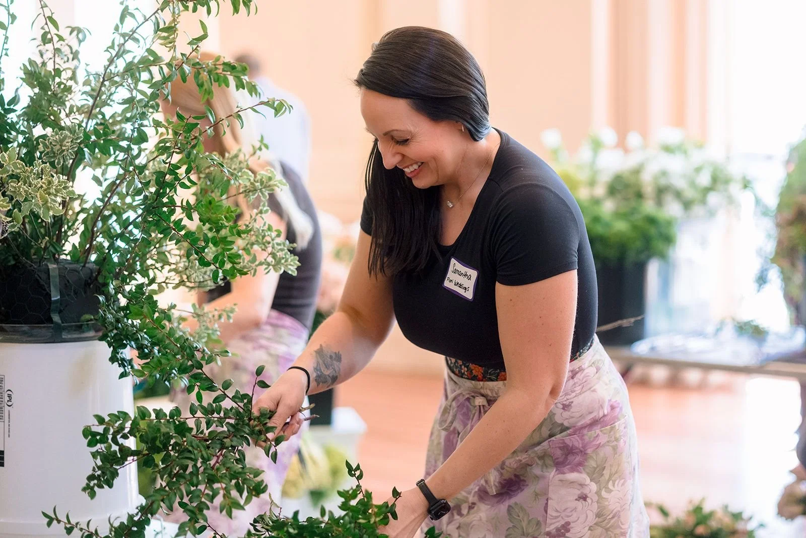 Wedding florist arranging a lush floral centerpiece with greenery, peach, and burgundy blooms, smiling while styling details for an elegant event.