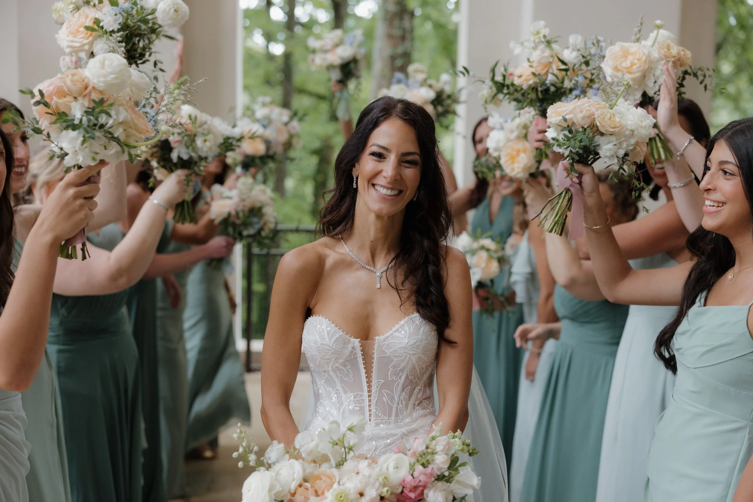 Smiling bride surrounded by bridesmaids raising ivory and blush bouquets in celebration