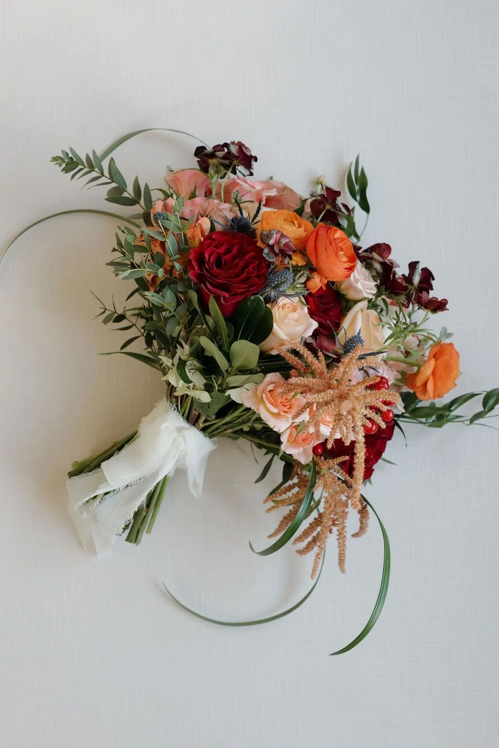Bridal bouquet of red, terracotta, and peach roses with trailing greenery and amaranthus, tied with white ribbon
