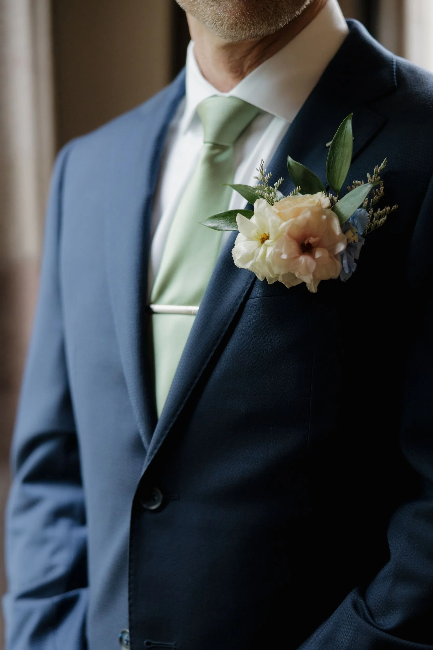 Close-up of a peach and blue boutonniere with greenery on a navy suit with a sage green tie