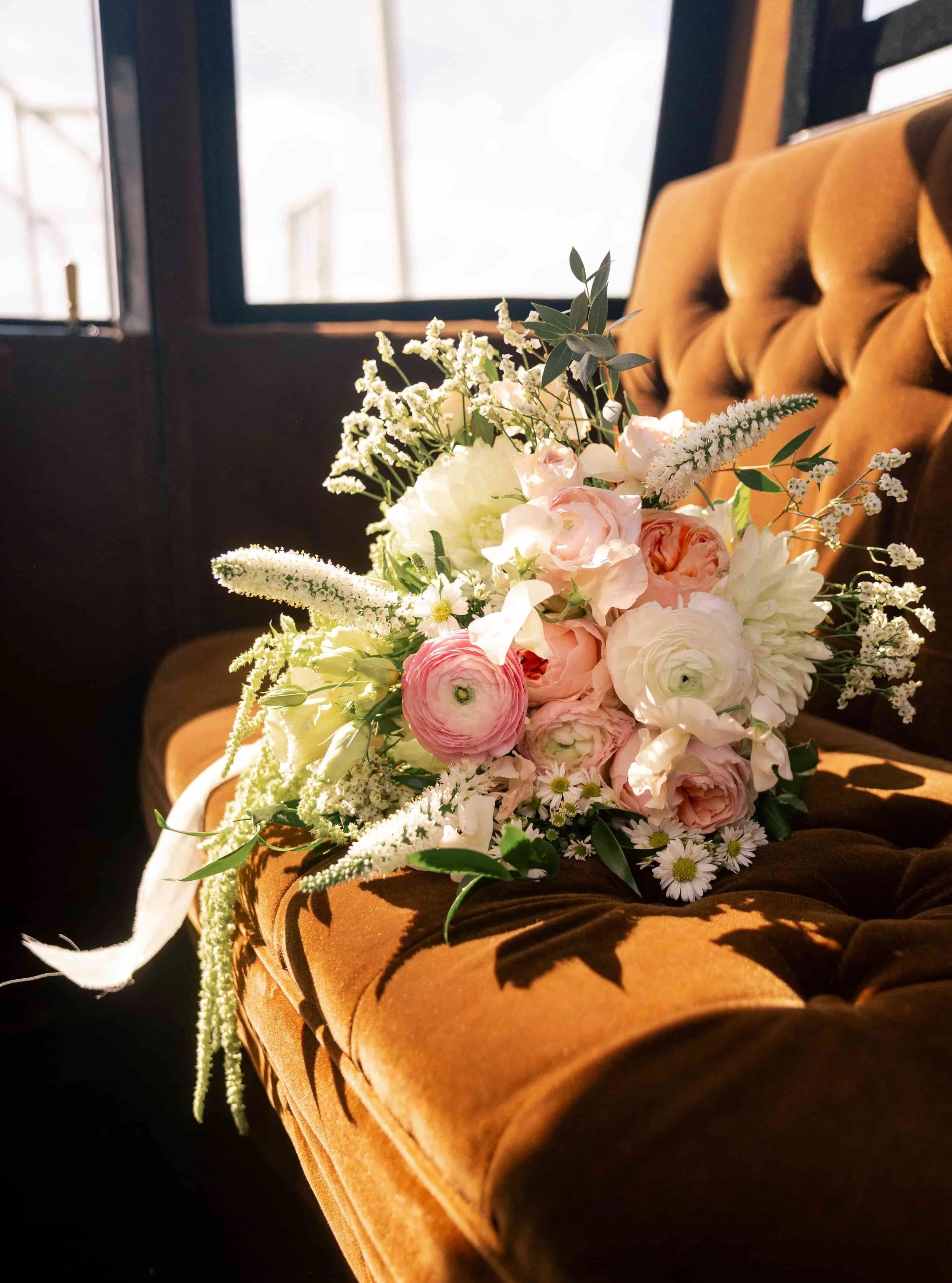 Bridal bouquet of blush and white ranunculus, roses, and wildflower accents resting on a vintage brown velvet seat, captured in warm natural light.