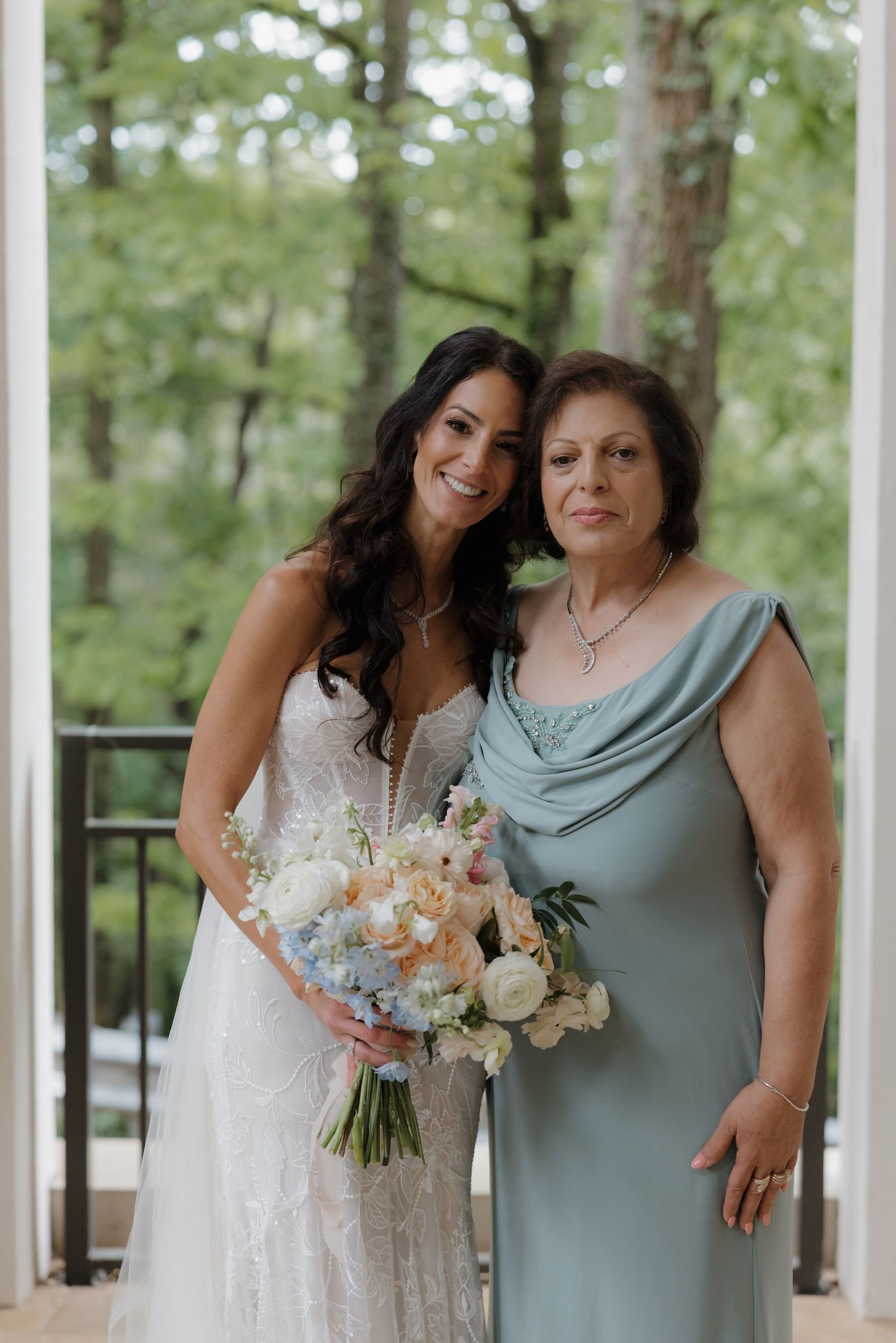 Bride holding her bouquet beside her mother in a sage gown on a garden veranda