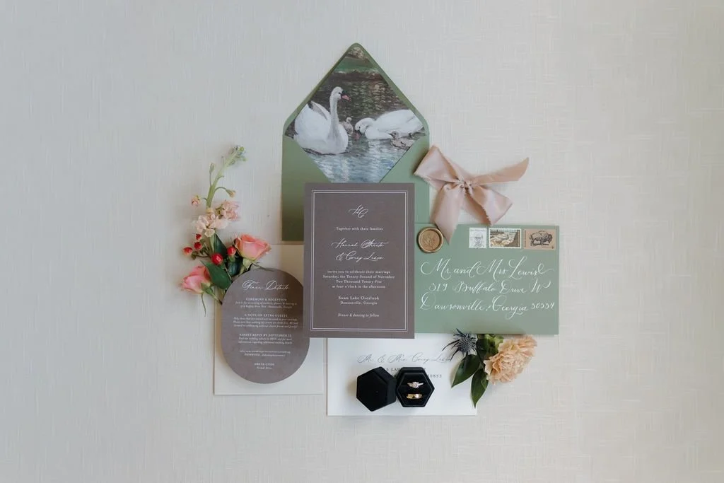 Wedding invitation suite flat lay with a swan-lined green envelope, blush ribbon, wax seal, ring box, and floral sprigs