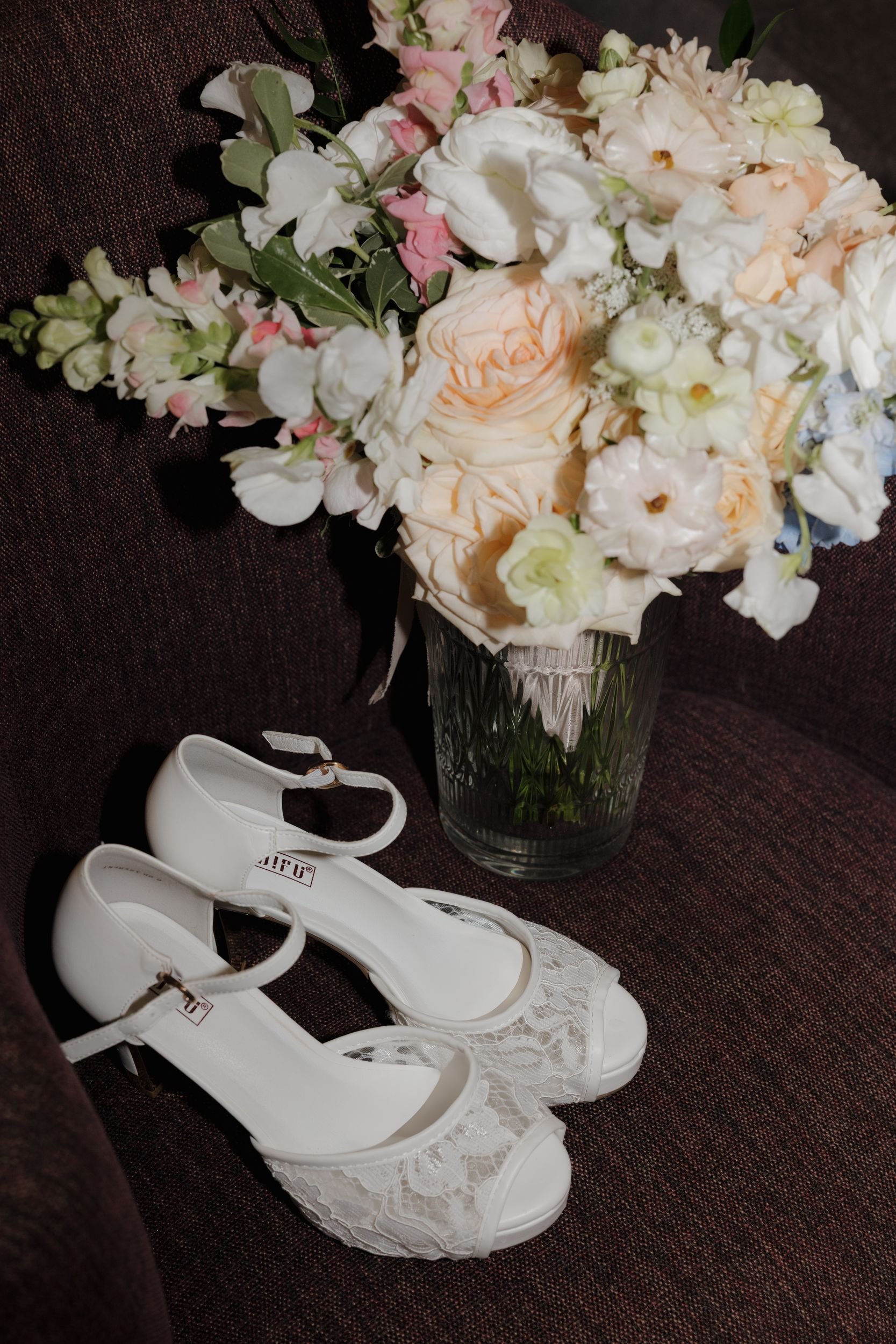 Bridal bouquet of peach and blush blooms in a glass vase beside white lace bridal heels