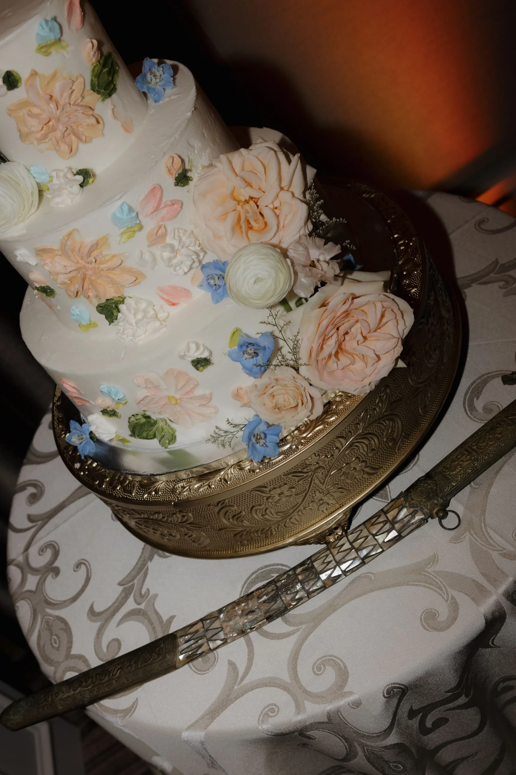White tiered cake with painted and fresh florals on an ornate gold stand with a serving knife