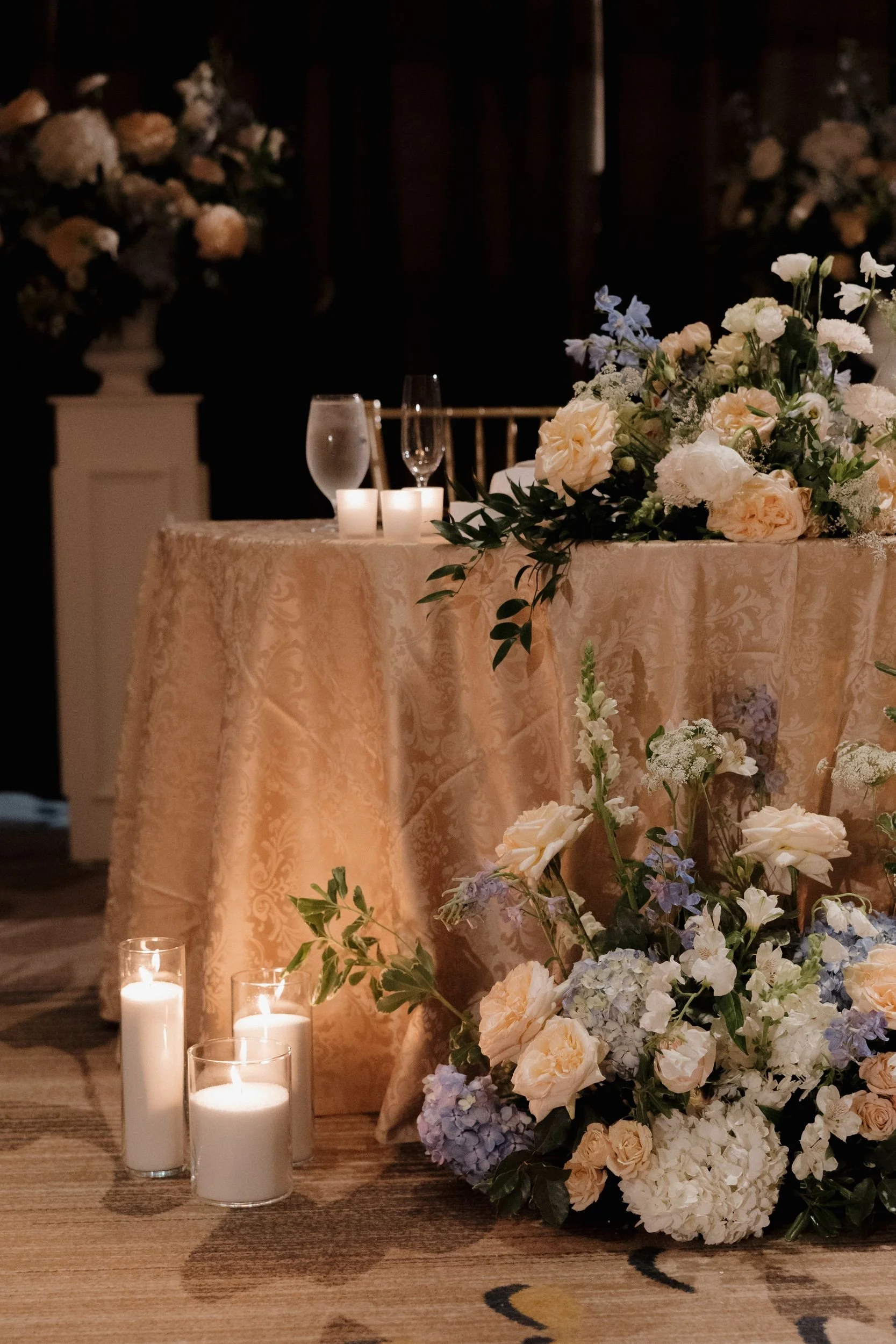 Draped sweetheart table with a cascading floral arrangement and a ground installation of blue hydrangea and white roses among pillar candles