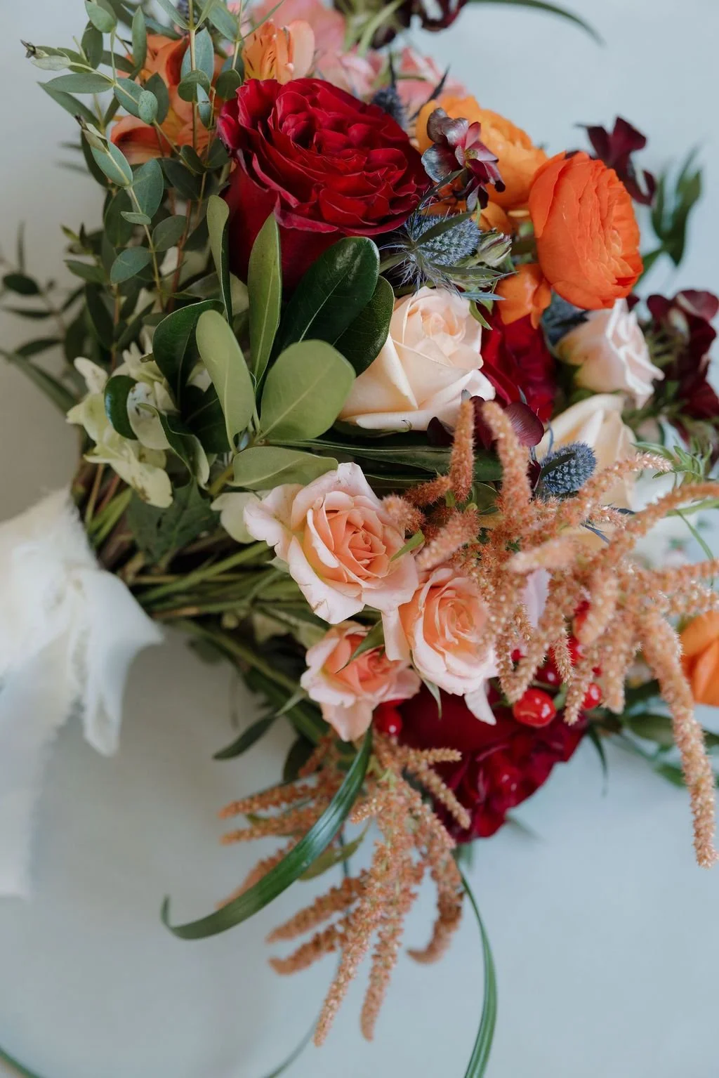 Close-up of a bouquet with red and peach roses, orange ranunculus, blue thistle, and trailing amaranthus