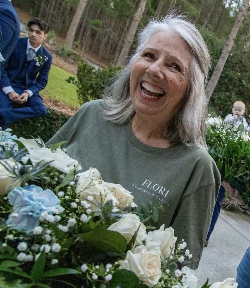 Wedding floral team member smiling while carrying bouquets of white roses, blue accents, and greenery on a wedding day.