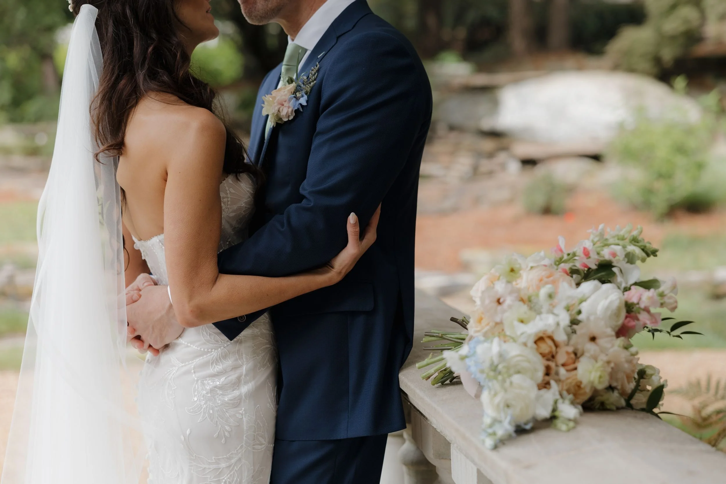 Bride and groom embracing outdoors, the bride's ivory and blush bouquet resting on a stone ledge