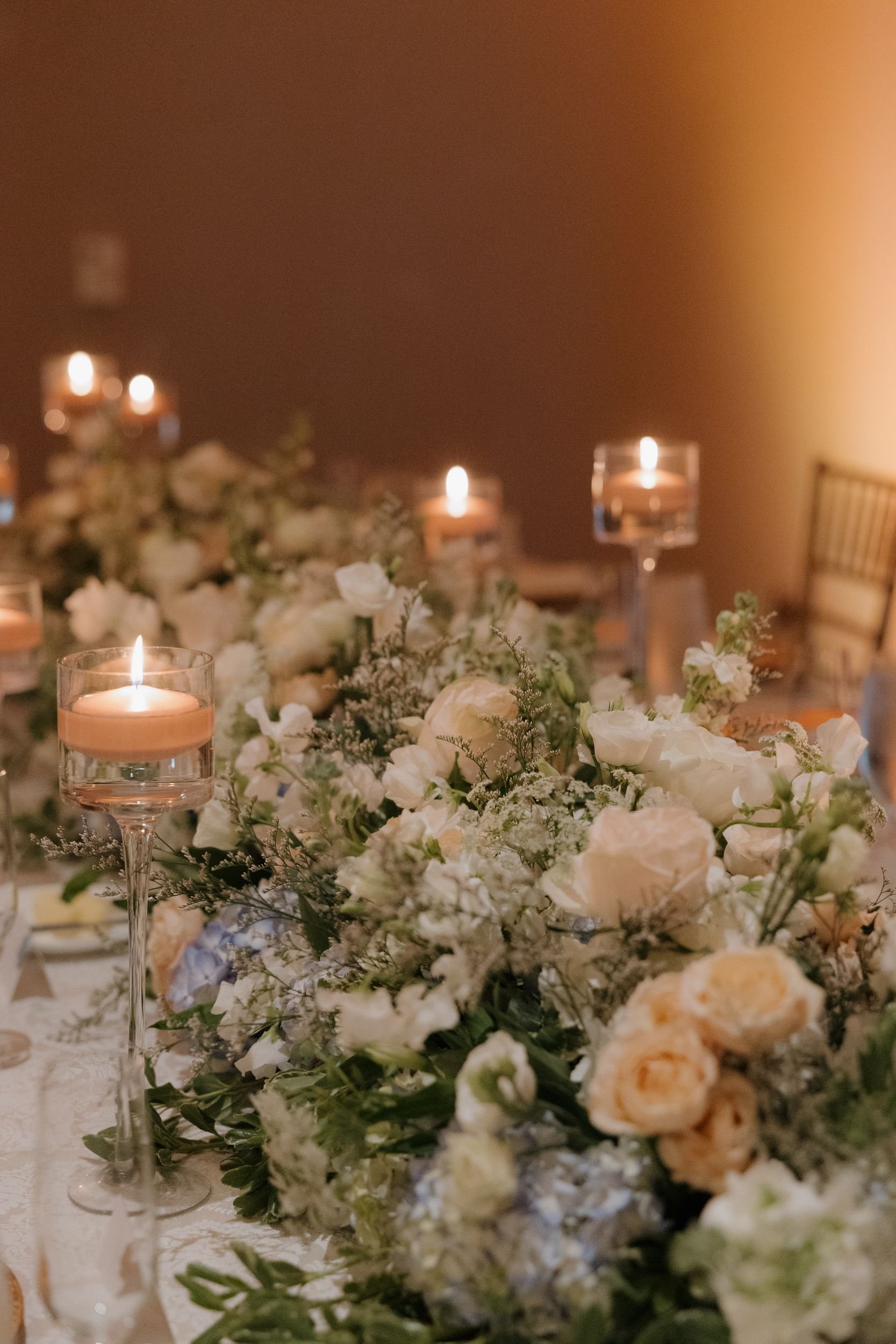 Close-up of a low table garland with white roses, blue hydrangea, and greenery among floating candles