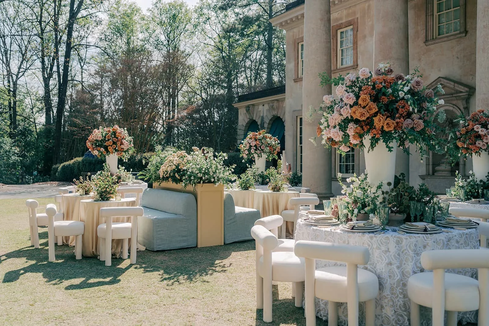 Elegant outdoor wedding reception at Swan House Atlanta featuring round tables with soft neutral linens, modern white chairs, and lush floral centerpieces in blush, peach, and greenery.