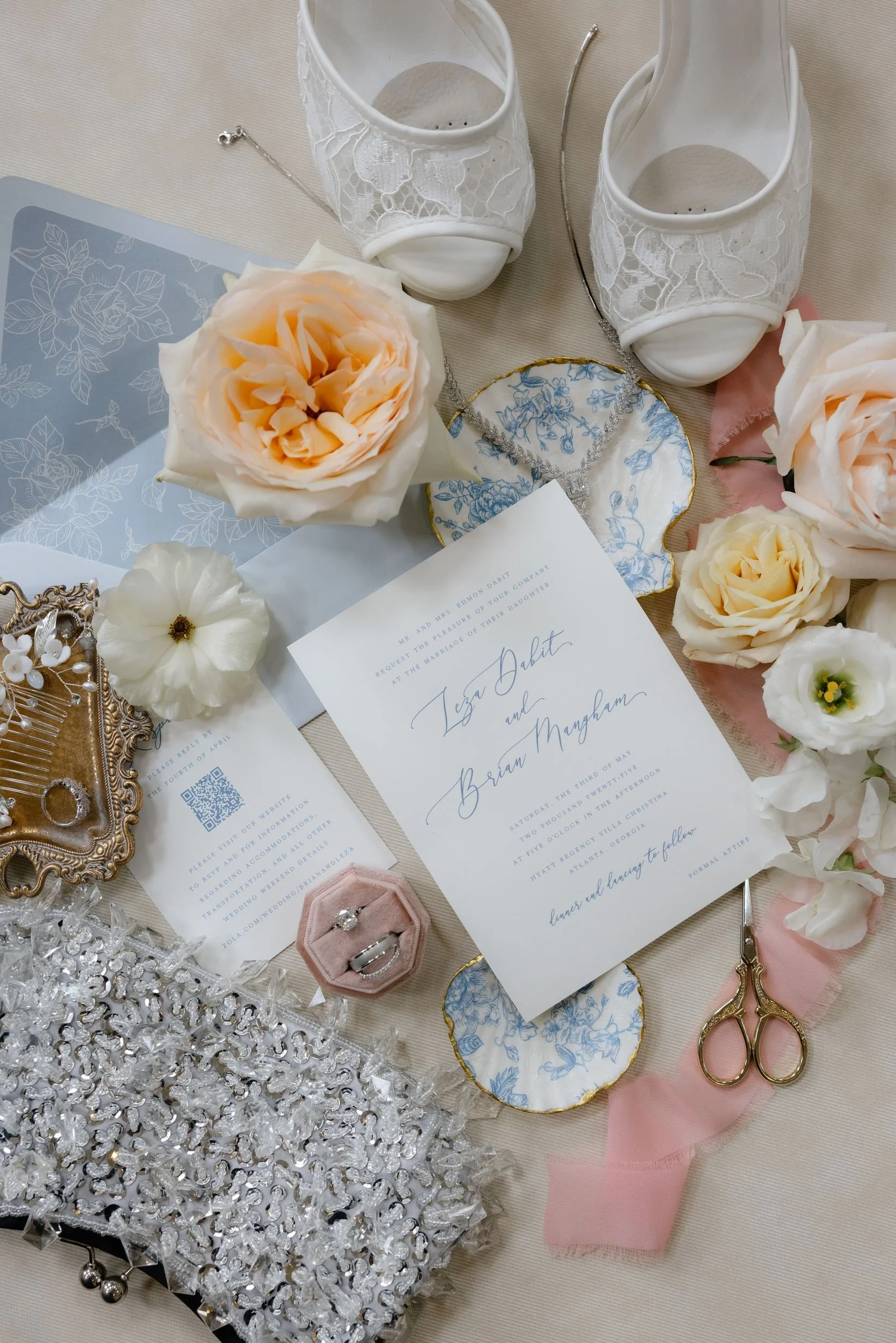 Wedding day flat lay with the invitation suite, peach garden roses, blue floral china, ring box, gold scissors, and bridal heels
