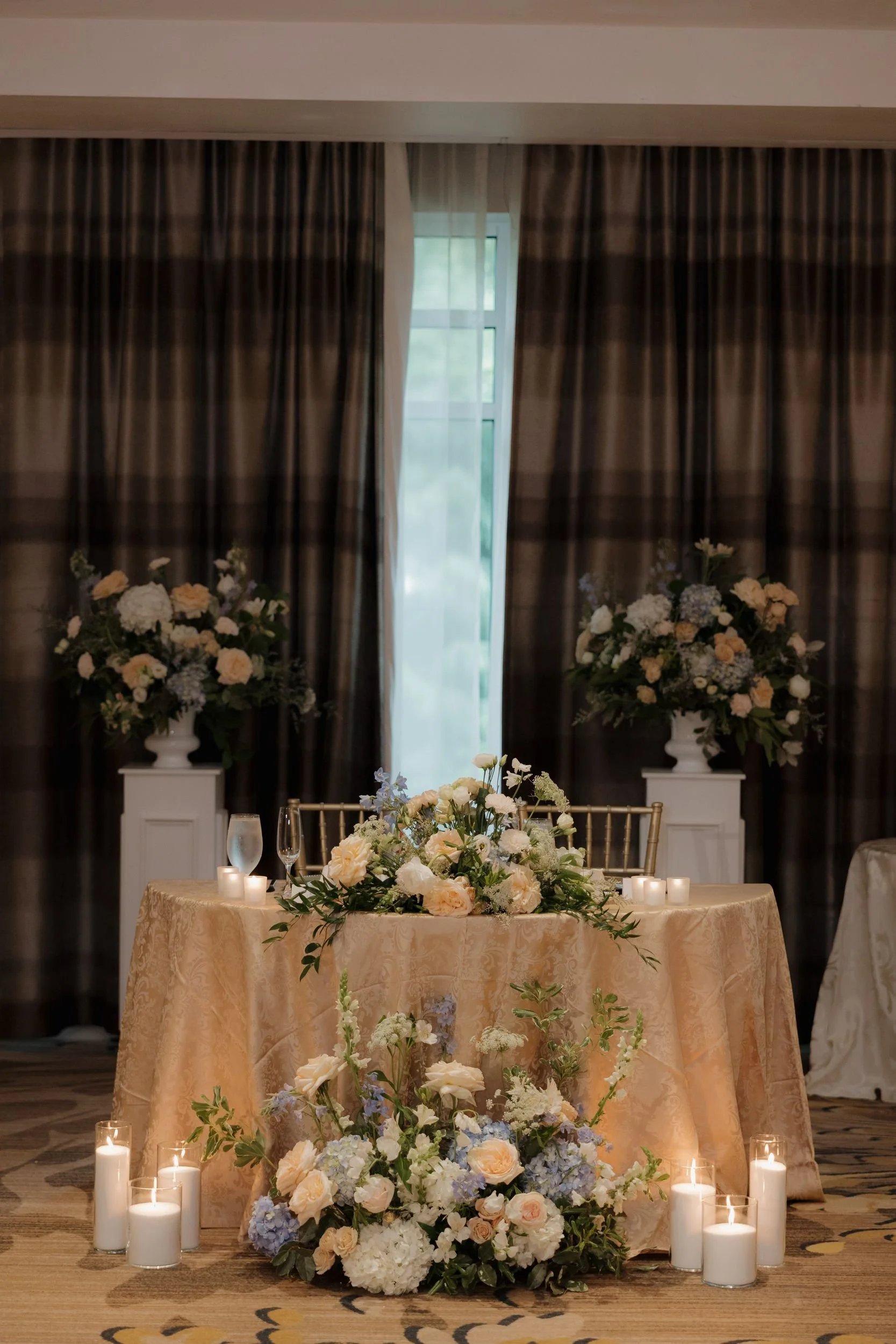 Sweetheart table draped in gold with a floral centerpiece, a ground installation of blue hydrangea and white blooms, candles, and flanking pedestal arrangements