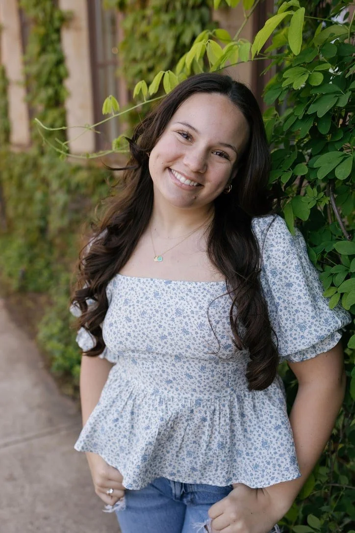 Smiling young floral designer standing outdoors against a wall of greenery, wearing a light blue floral blouse and jeans.
