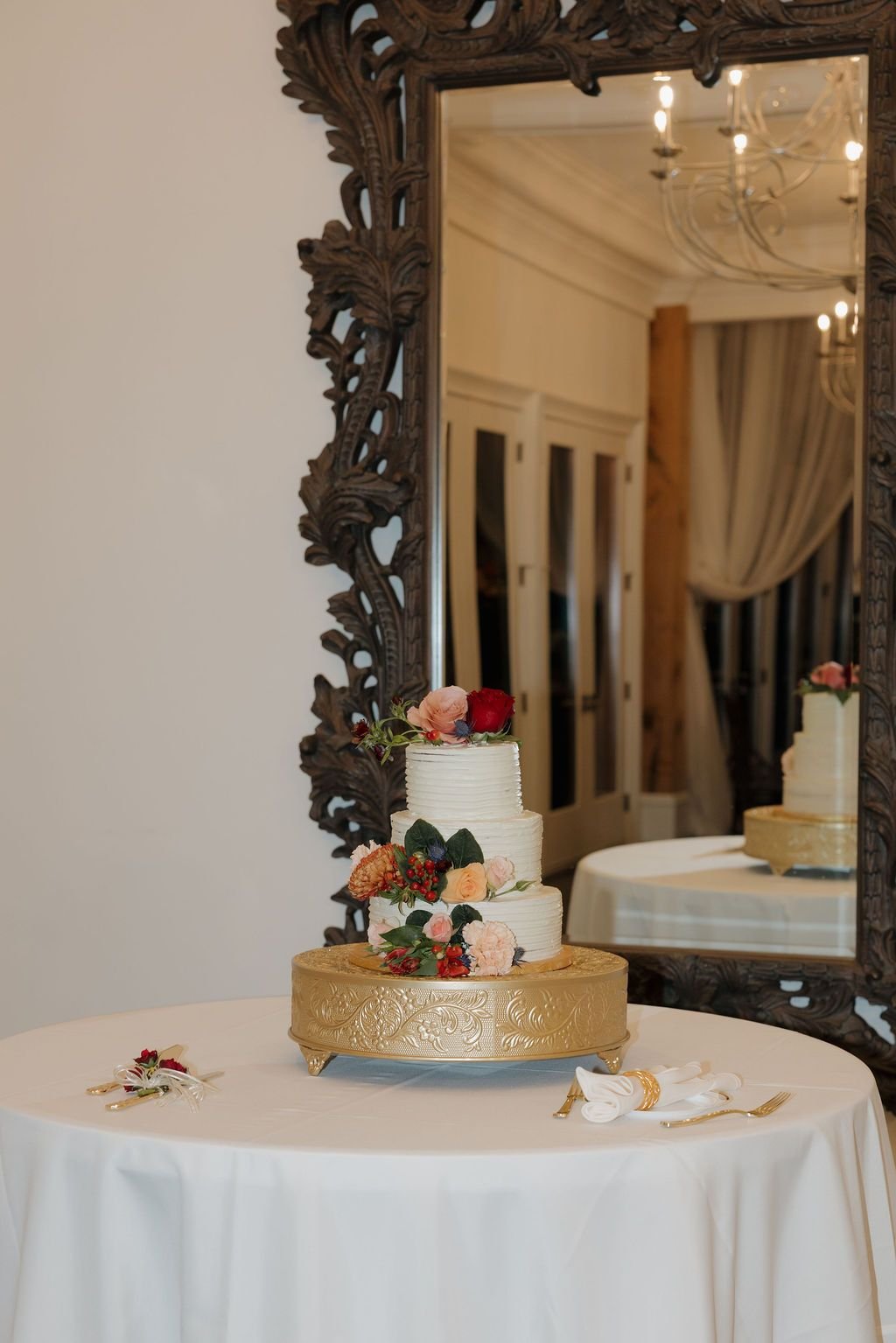 Three-tier white wedding cake on a gold stand accented with peach and burgundy florals before an ornate mirror