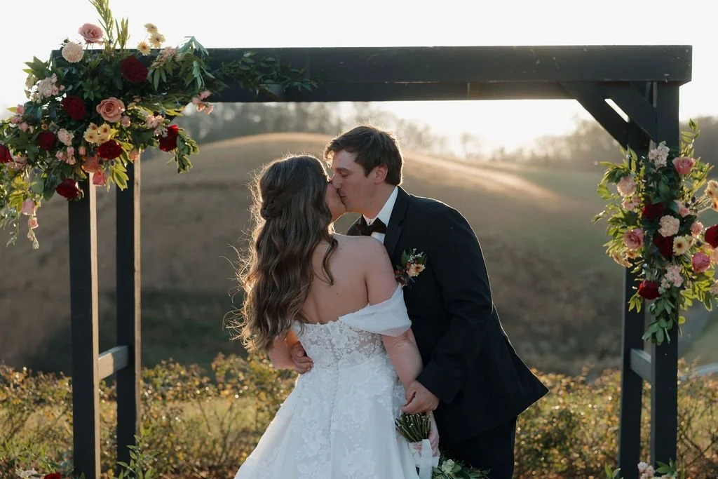 Couple kissing beneath a wooden ceremony arbor decorated with burgundy and peach floral clusters at golden hour