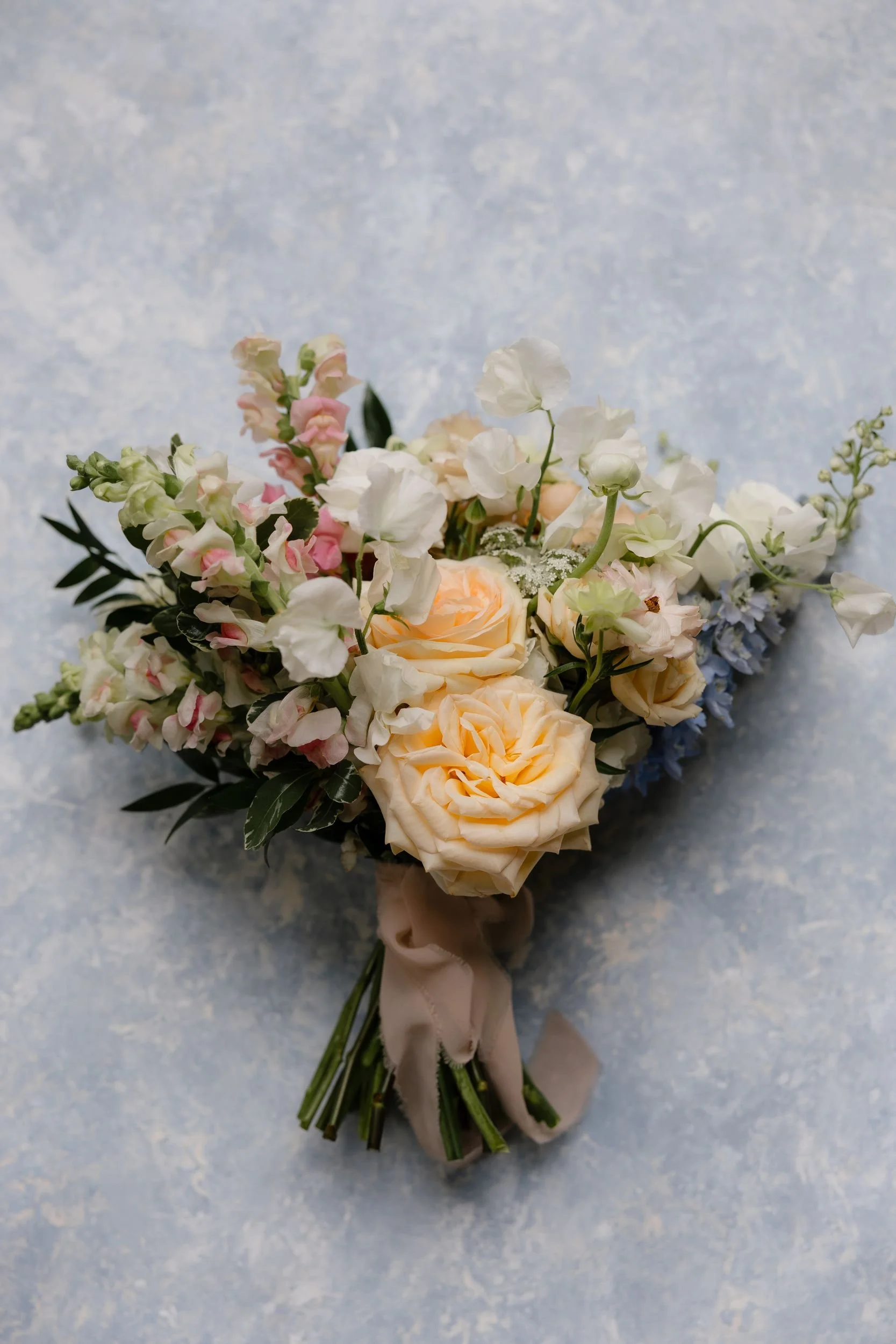 Bridal bouquet of peach garden roses, blush snapdragons, blue delphinium, and greenery tied with ribbon on a marbled surface