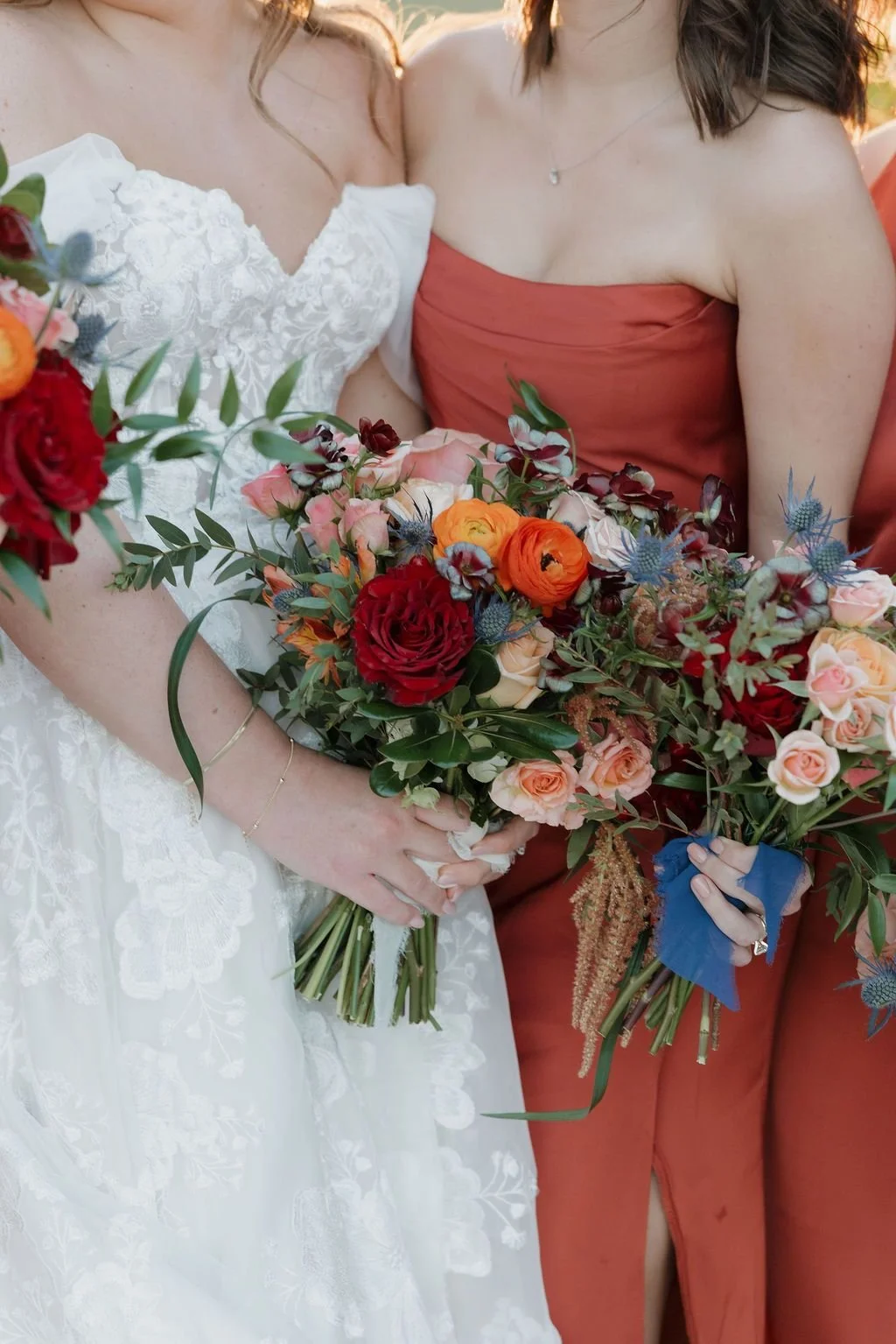 Bride and bridesmaid holding bouquets of red roses, orange ranunculus, blue thistle, and amaranthus
