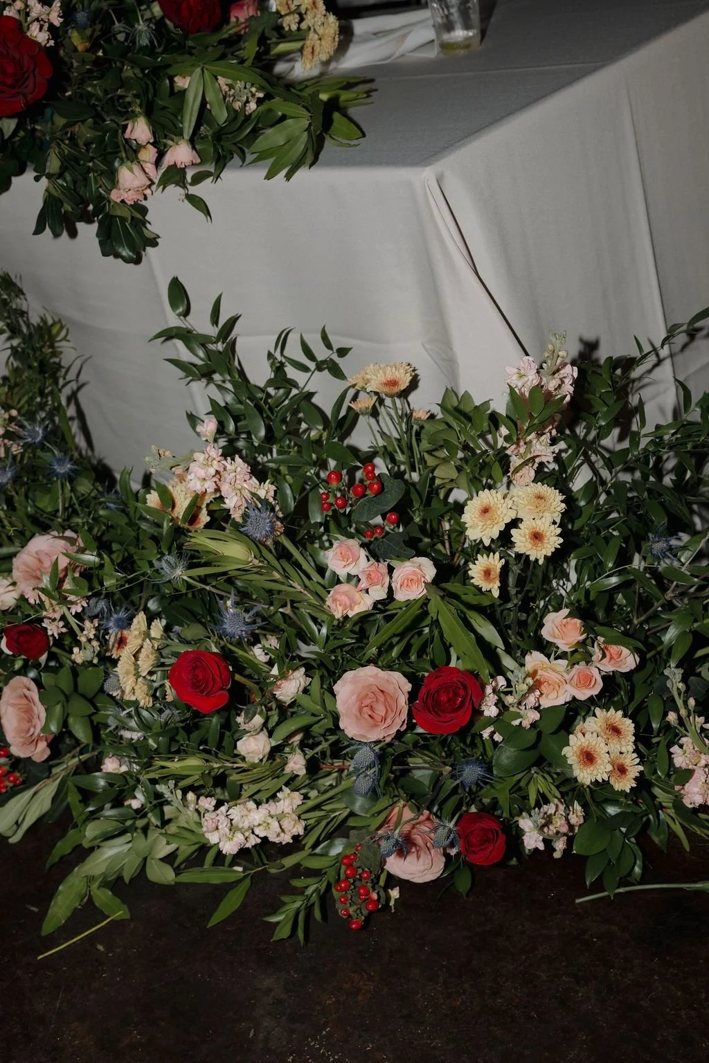 Lush ground floral installation of red and peach roses, white dahlias, and greenery at the base of a draped table