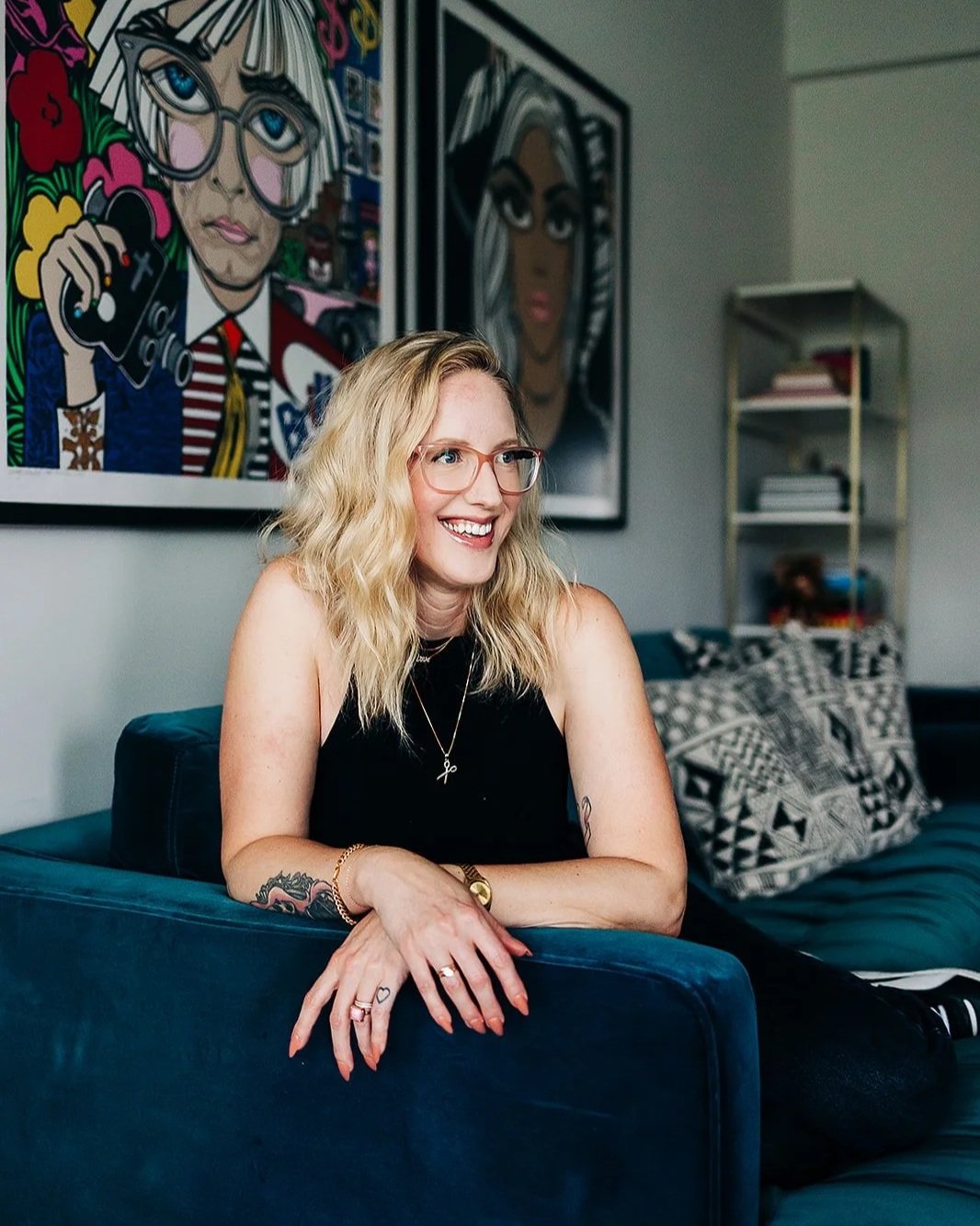 A woman with blonde wavy hair, wearing glasses and a black sleeveless top, sits on a blue couch, smiling. Behind her are two colorful art portraits on the wall.