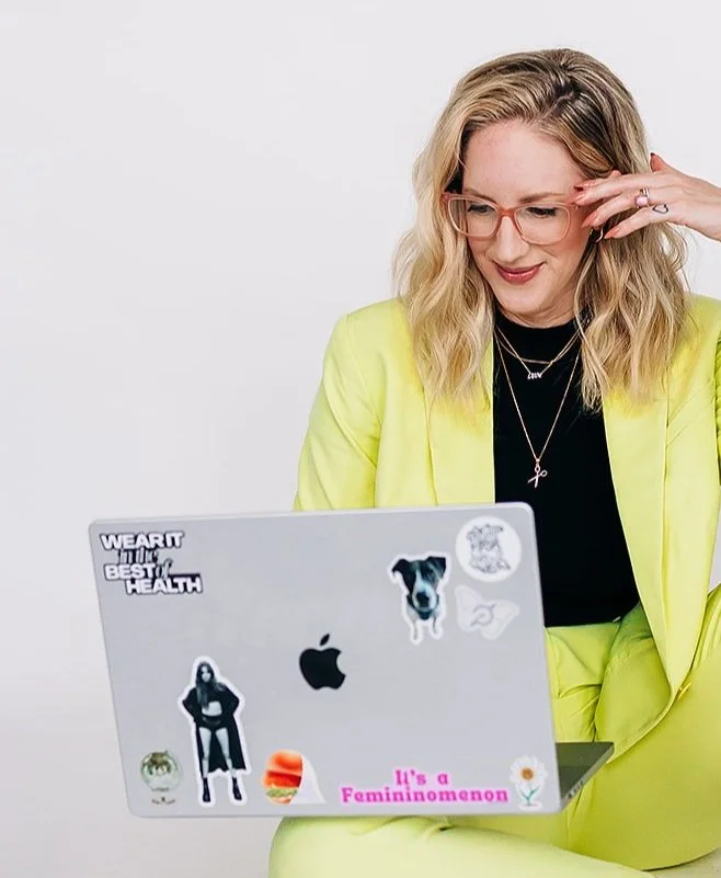 Amy Moscowitz with blonde hair, pink glasses, and a yellow blazer working on a laptop decorated with stickers, with a plain white background.