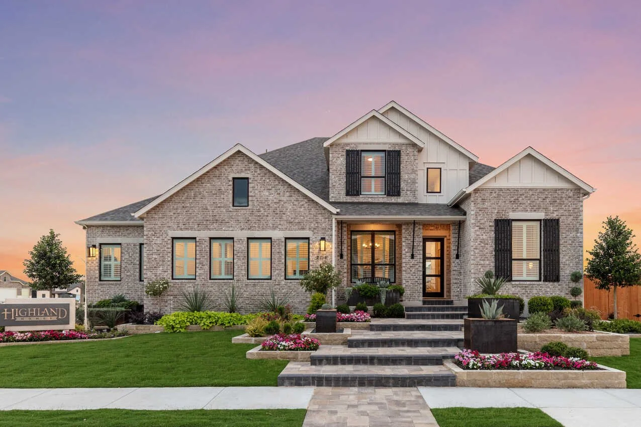 A modern two-story brick house with a landscaped front yard at sunset, featuring a walkway, stairs, and flower beds.