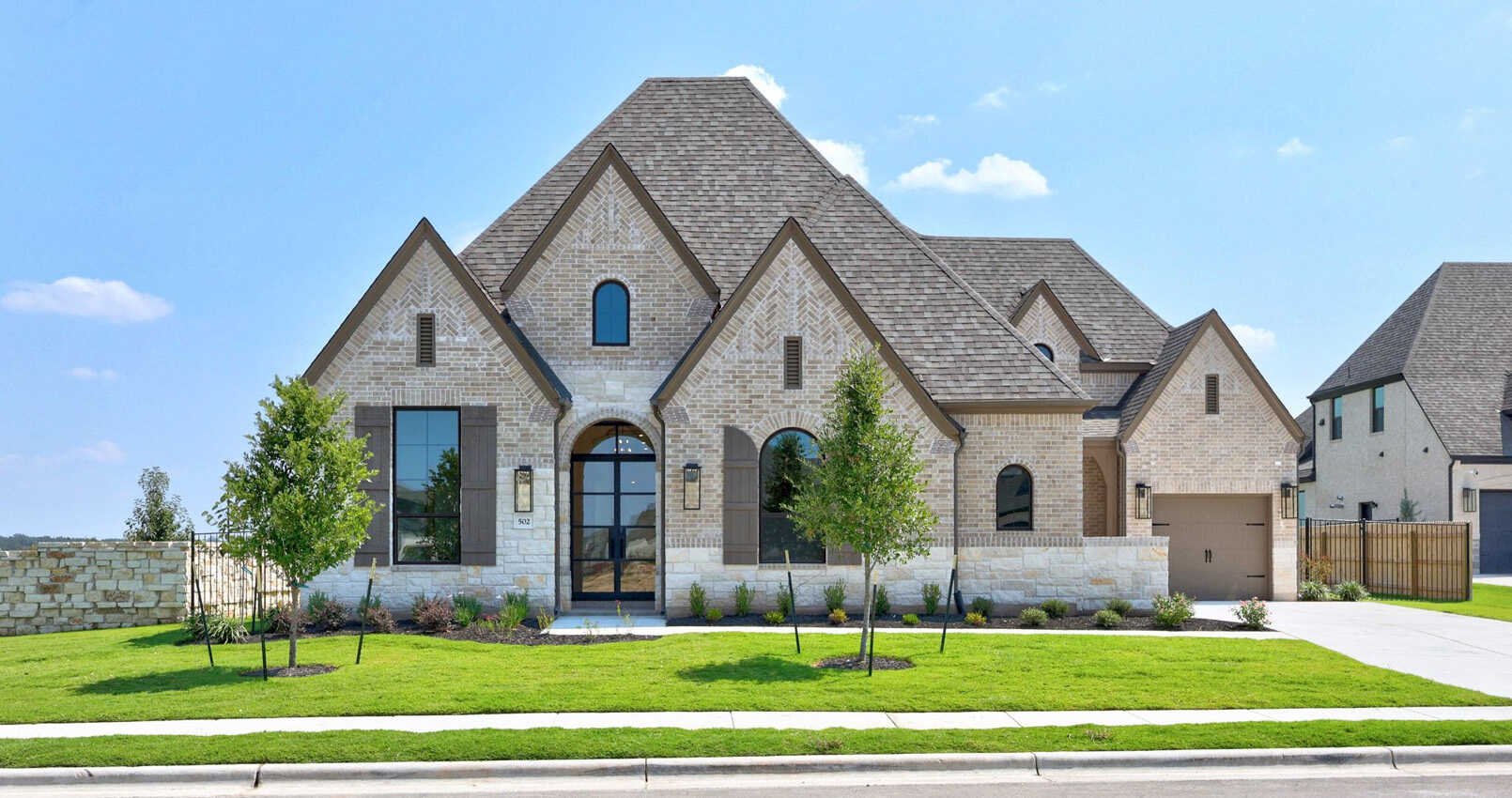 A large, modern suburban house with brick and stone exterior, front yard with green grass, trees, and sidewalk under a blue sky.