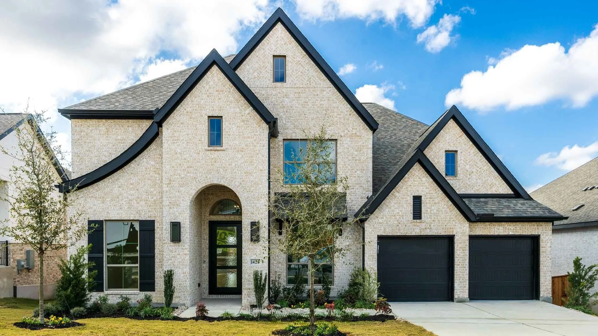 Modern two-story house with light brick exterior, black trim, and a two-car garage, surrounded by a landscaped yard with small trees and plants, under a blue sky with clouds.