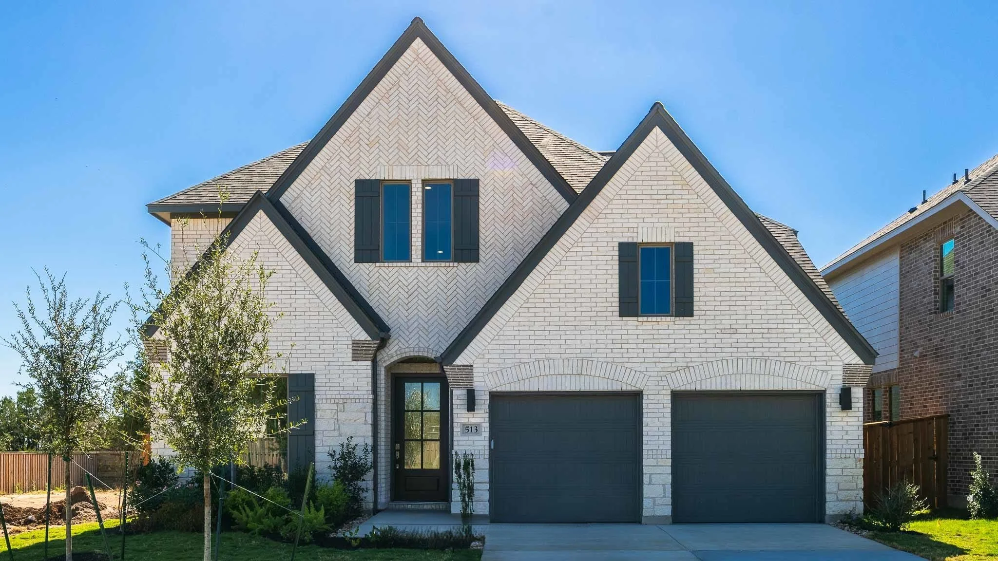 Modern two-story house with white brick exterior, dark shutters, and a double garage with dark doors, set against a blue sky.