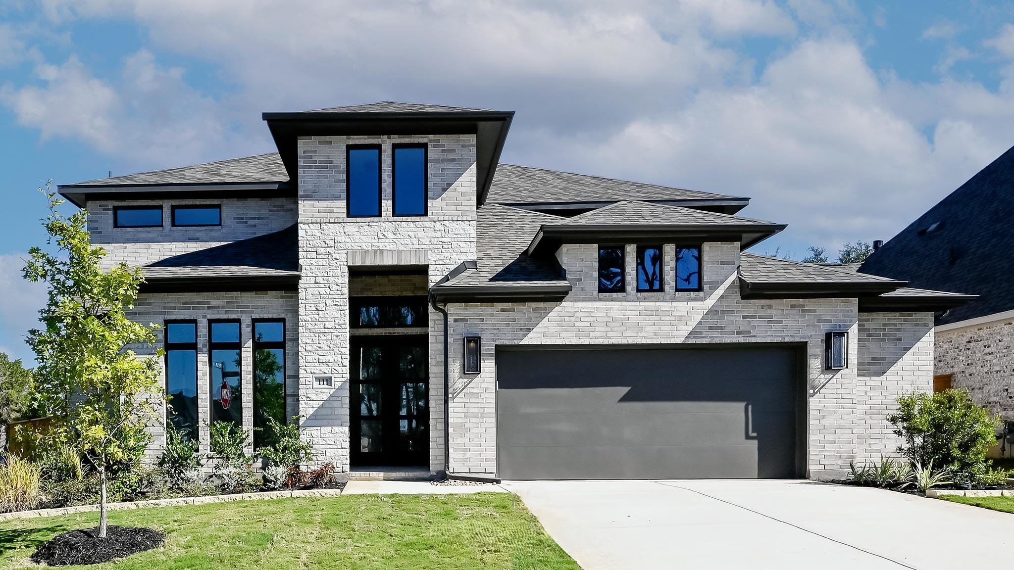 Modern two-story house with gray and white brick exterior, black-framed windows, and a gray garage door, with a small front lawn and trees.