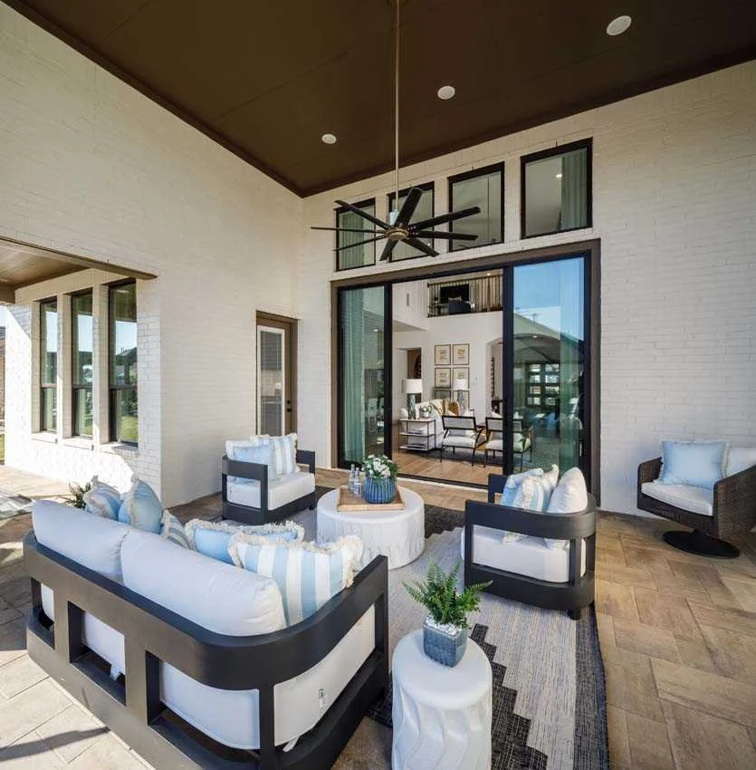 Covered outdoor patio with white brick walls, black ceiling fan, glass doors, and outdoor seating including white and black furniture with striped pillows, a round coffee table, potted plant, and connected indoor living room visible through glass doors.