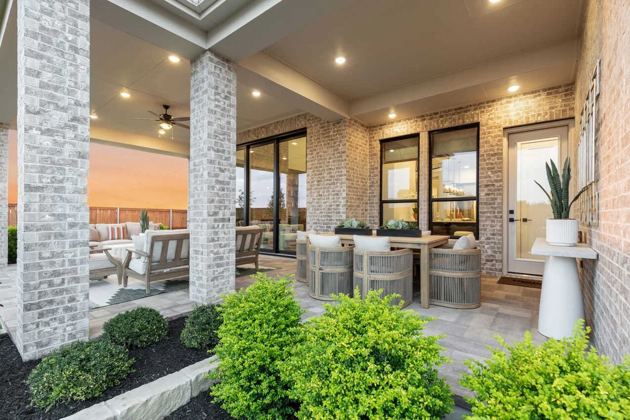 Covered outdoor patio with seating area, brick walls, large windows, potted plants, and greenery in foreground, at sunset.