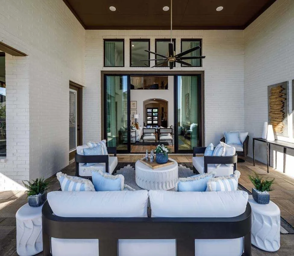An indoor-outdoor living space with white brick walls, black-framed windows, a ceiling fan, and a modern seating area with white sofas and striped pillows, a coffee table with decor, and potted plants.