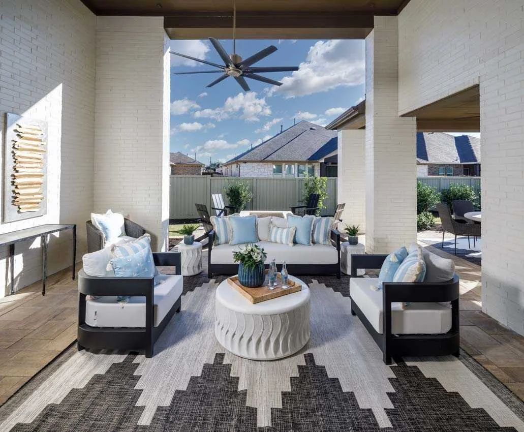 Covered outdoor patio with white brick walls, ceiling fan, modern seating with blue and white pillows, a round white coffee table with a plant and drinks, and a gray patterned rug. Clear sky with clouds visible.