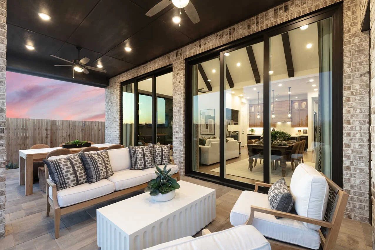 An outdoor patio with a white cushioned sofa and chairs, a white coffee table with a potted plant, a dining table with chairs, and a view into a brightly lit indoor living and dining area through large glass sliding doors. The patio has a brick wall 