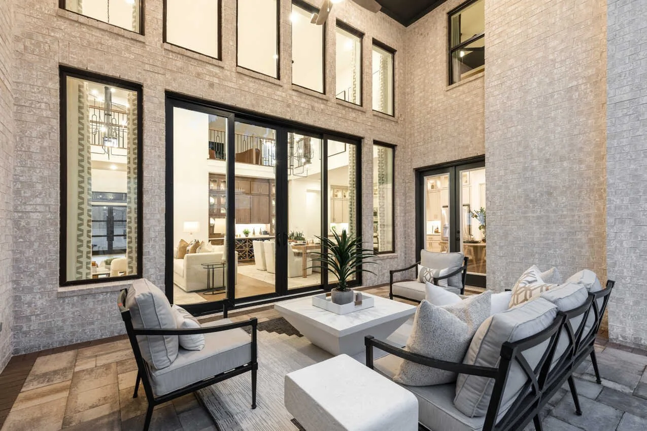 Outdoor patio with seating area featuring white cushioned chairs and sofa, a white coffee table with a plant, enclosed by a brick wall with large glass doors and windows revealing an interior living space.