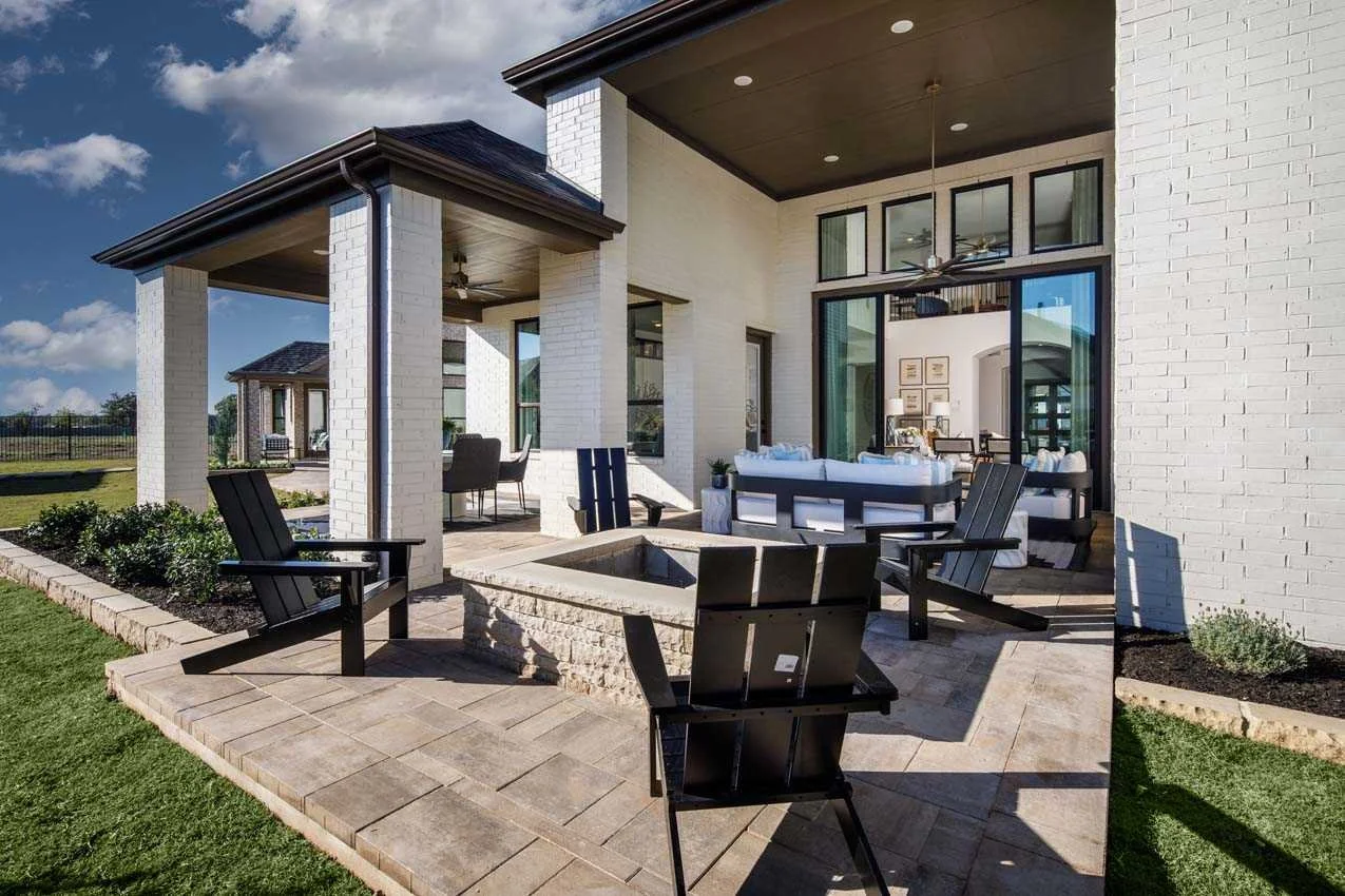 Modern outdoor patio with black Adirondack chairs, a fire pit, a white brick house with large glass windows, and a landscaped yard under a partly cloudy sky.