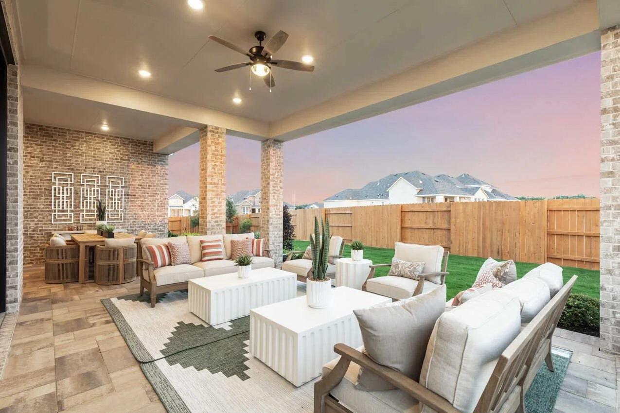 Covered outdoor patio with beige couches and chairs, white coffee tables, potted plants, brick walls, ceiling fan, and a view of a neighborhood with houses and a pinkish sky in the background.