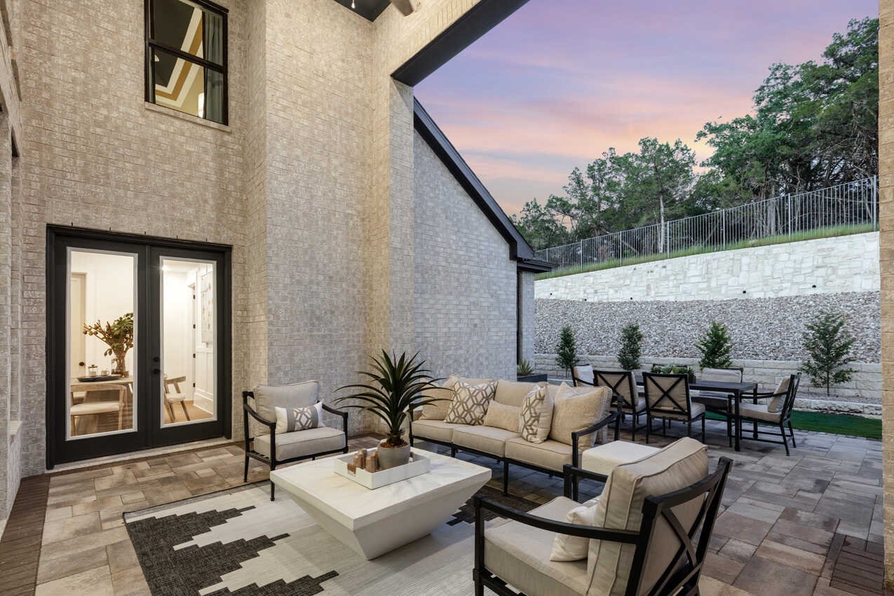 Outdoor patio with beige brick walls, modern black framed glass doors, and gray tiled flooring. Furniture includes a white sofa, armchairs, and a coffee table with a plant. In the background, there are dining chairs and a table with a grassy area and