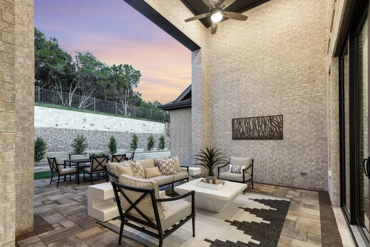 Covered outdoor patio with seating including a sofa and chairs, a coffee table with a potted plant, a black and white rug, and a ceiling fan, overlooking a backyard with a hillside, trees, and a sunset sky.