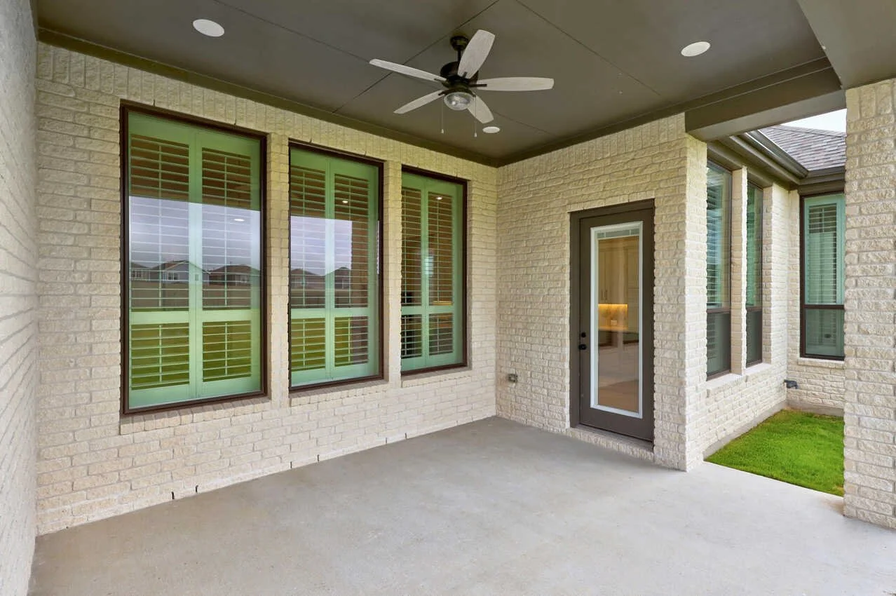 A covered patio with a ceiling fan, beige brick walls, large windows with green shutters, a glass door, and a small patch of grass outside.
