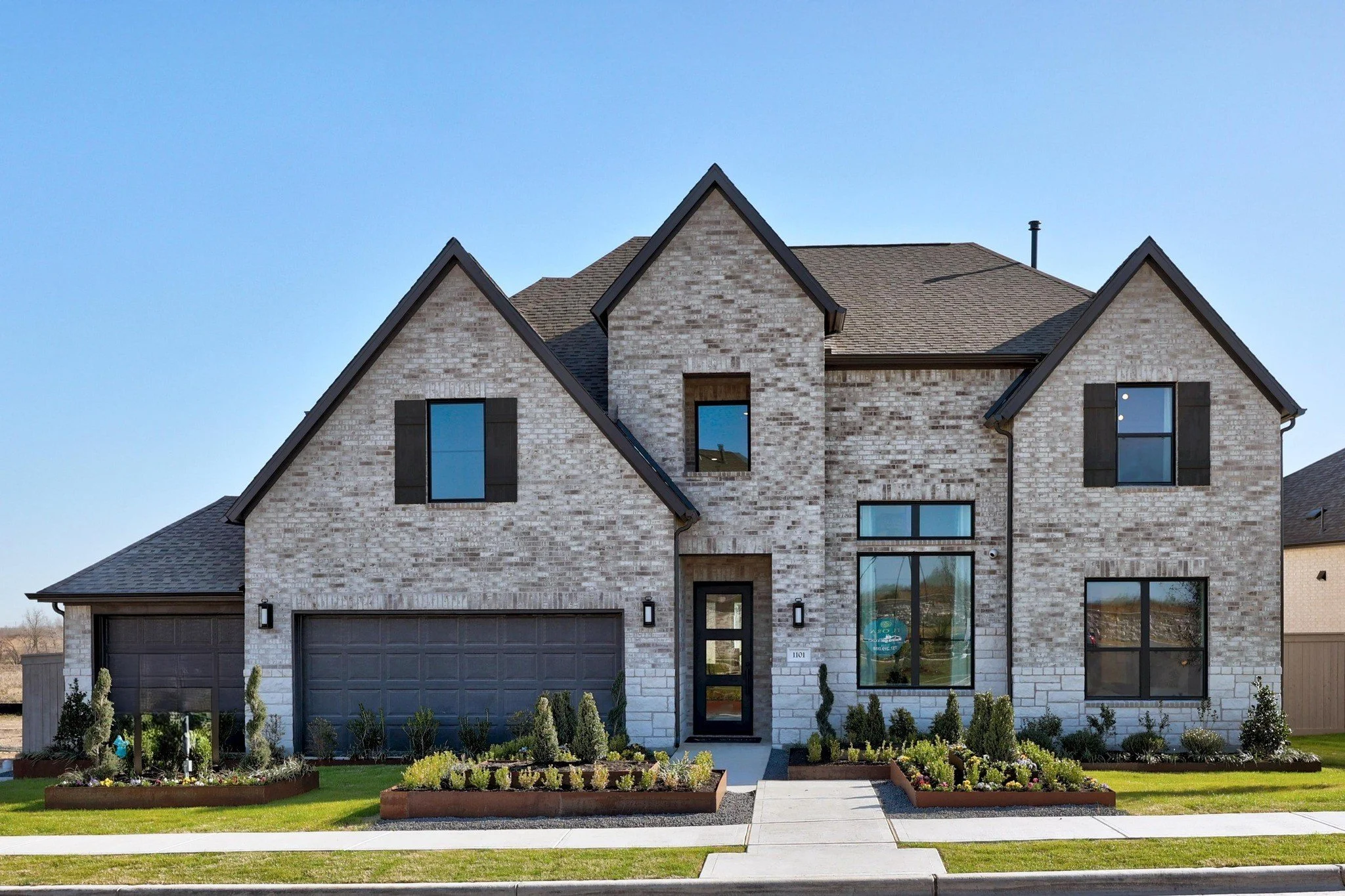 Front view of a modern two-story brick house with a dark gray garage door and black fixtures, surrounded by manicured lawns and planting beds with small shrubs and flowers.