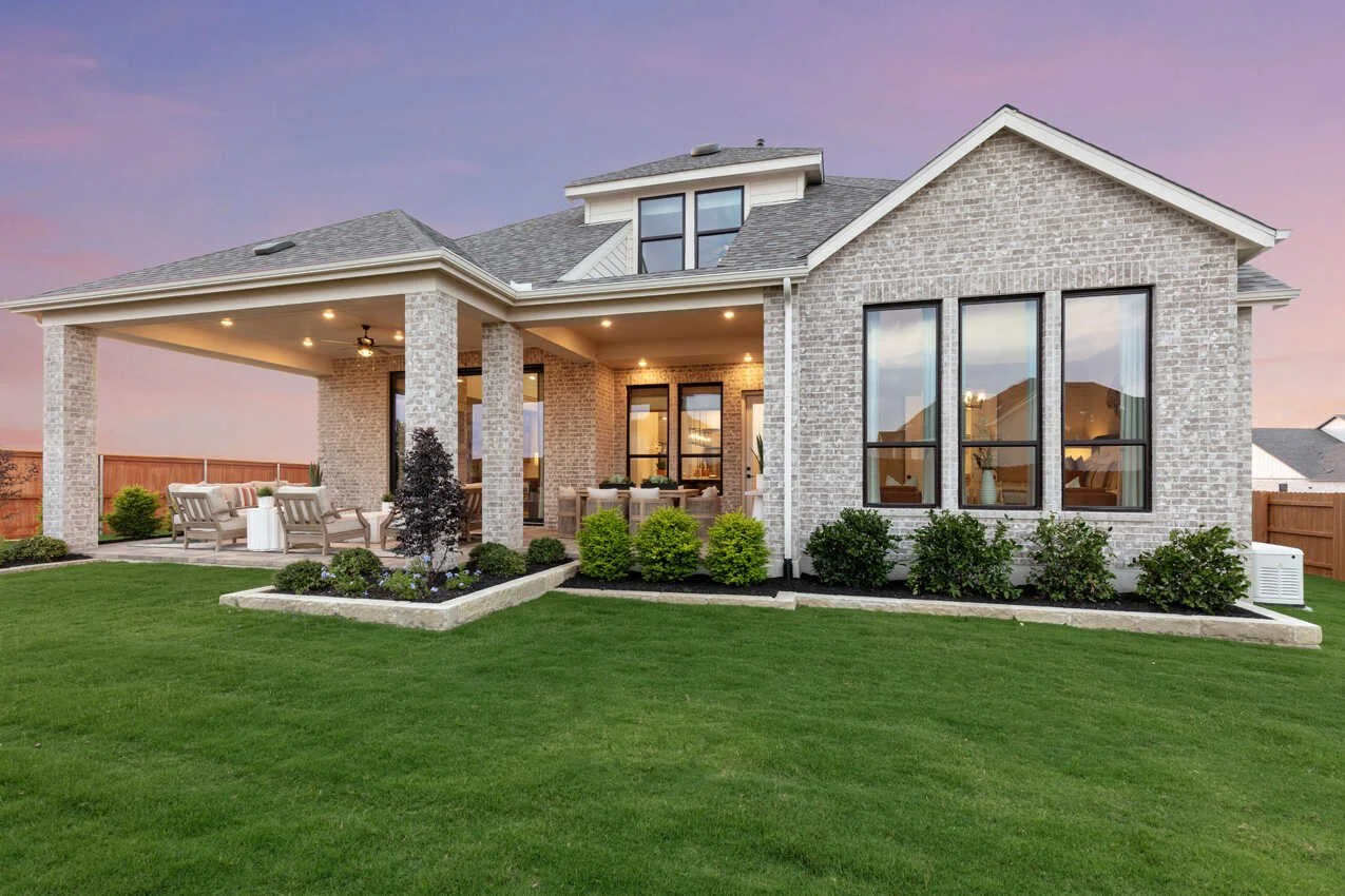 Modern brick house with large windows and a spacious backyard patio with outdoor furniture, surrounded by a well-manicured lawn and garden, during sunset.